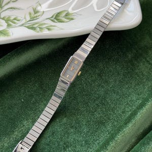 Puede incluir: Un reloj de pulsera rectangular con una correa de metal plateado y una esfera gris. La esfera del reloj tiene detalles dorados y números romanos. El reloj se muestra sobre una superficie de terciopelo verde.