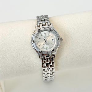 May include: A silver-toned Wrangler quartz watch with a round face and a date display at the 3 o'clock position. The watch has a silver band and a silver dial with black numerals. The watch is water resistant.