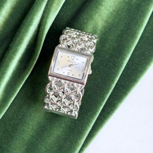 May include: A silver-tone wristwatch featuring a square face with a white dial, black numerals at the 12, 3, and 6 positions, and the text STUDIO. The watch has a decorative, interlocking chain-link bracelet band and a quartz movement.
