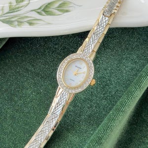 May include: A gold and silver-toned SHARP quartz watch with a round face. The watch face is surrounded by a ring of small, clear crystals. The watch band is a combination of gold and silver links. The watch is displayed on a green surface.