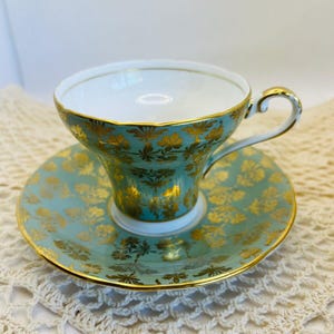 May include: A vintage teacup and saucer set. The cup and saucer are a light teal color with a gold floral pattern and gold trim. The teacup has a curved handle and a white interior. The saucer is round and has a matching design.