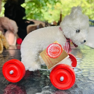 Puede incluir: Un oso de peluche blanco vintage con ruedas rojas. El oso tiene una cinta a rayas rojas y blancas alrededor del cuello y una etiqueta que dice "Teddy Original". El juguete está hecho de material de felpa.