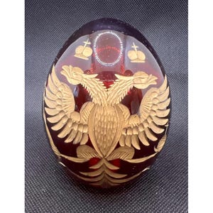 Può includere: Un uovo di vetro rosso intenso con un emblema inciso di un'aquila bicipite color oro centrato sulla parte anteriore. Sopra l'aquila ci sono due piccole corone incise. La superficie del vetro è liscia e riflettente.