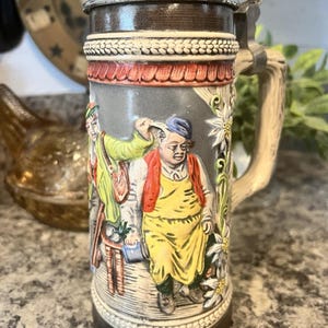 May include: A ceramic beer stein featuring a metal lid and a handle. The body displays a relief scene of two figures, one in a yellow apron and one in a green jacket, surrounded by white edelweiss flowers against a gray background.