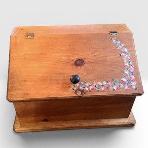 Peut inclure: Une boîte de rangement rectangulaire en bois avec un couvercle incliné à charnières. Le bois naturel brun clair présente un bouton circulaire sombre et un motif floral peint à la main en rose, bleu et vert sur le côté droit et le bord inférieur.