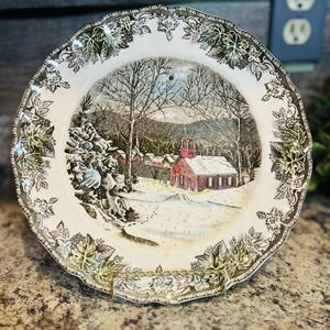 May include: A vintage decorative plate with a winter scene. The plate features a red church in a snowy landscape, surrounded by trees and a decorative leaf border. The plate has a scalloped edge.