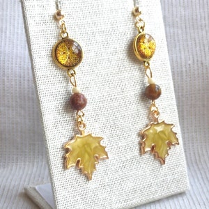 May include: A pair of gold-toned earrings with a fall theme. Each earring features a round, amber-colored charm with a floral design, a brown bead, and a yellow maple leaf charm. The earrings are displayed on a white stand.