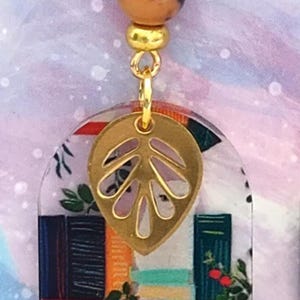 May include: A pair of dangle earrings featuring a gold-colored leaf charm and a clear, arched pendant with a cat and bookshelf design. The earrings have gold-colored beads and accents.