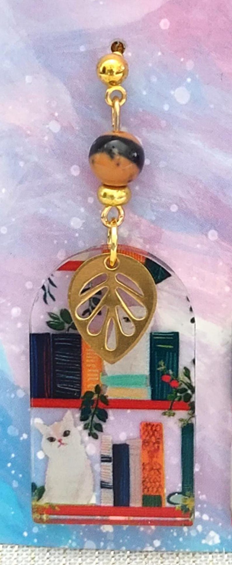 May include: A pair of dangle earrings featuring a gold-colored leaf charm and a clear, arched pendant with a cat and bookshelf design. The earrings have gold-colored beads and accents.