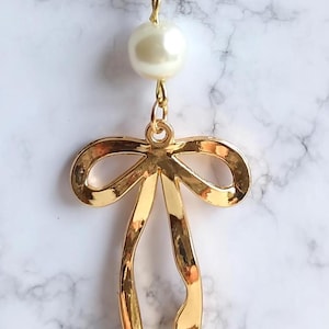 May include: Gold-toned necklace featuring a large, glossy faux pearl and a bow pendant. The bow pendant has a polished finish and long, flowing ribbons. The necklace hangs on a delicate gold chain, creating an elegant accessory.