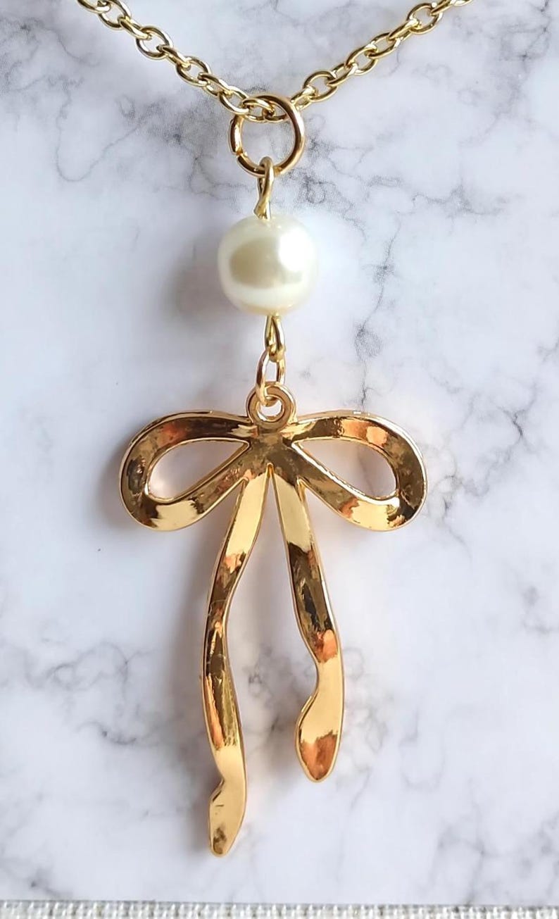 May include: Gold-toned necklace featuring a large, glossy faux pearl and a bow pendant. The bow pendant has a polished finish and long, flowing ribbons. The necklace hangs on a delicate gold chain, creating an elegant accessory.