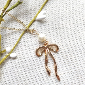 May include: A gold-colored necklace with a bow pendant and a pearl accent. The bow pendant has a looped design with flowing ends. The necklace is displayed on a textured, off-white surface, with sprigs of greenery and white floral accents.