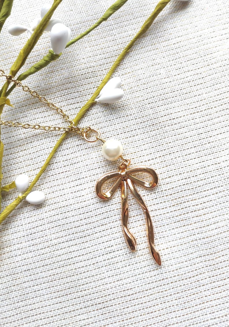 May include: A gold-colored necklace with a bow pendant and a pearl accent. The bow pendant has a looped design with flowing ends. The necklace is displayed on a textured, off-white surface, with sprigs of greenery and white floral accents.