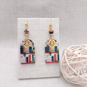 May include: A pair of dangle earrings featuring a whimsical design. Each earring has a gold-colored leaf charm, a patterned rectangular pendant with a cat and books, and a bead. The earrings are suspended from gold-colored ear wires.