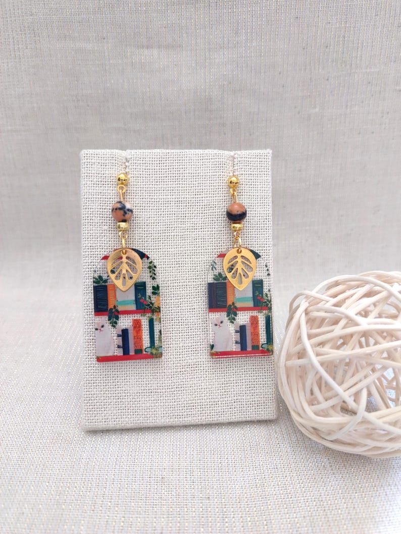 May include: A pair of dangle earrings featuring a whimsical design. Each earring has a gold-colored leaf charm, a patterned rectangular pendant with a cat and books, and a bead. The earrings are suspended from gold-colored ear wires.