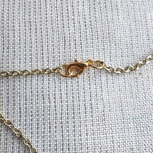 May include: Close-up of a gold-colored necklace chain with a lobster clasp. The chain is made of small, linked segments. The clasp is attached to the chain, ready to be fastened. The background is a textured, light-colored fabric.