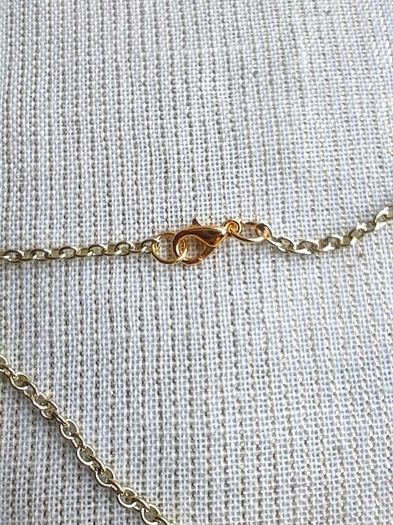 May include: Close-up of a gold-colored necklace chain with a lobster clasp. The chain is made of small, linked segments. The clasp is attached to the chain, ready to be fastened. The background is a textured, light-colored fabric.