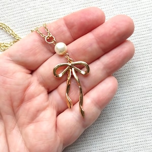 May include: A gold-colored necklace featuring a bow pendant with a dangling design. The necklace includes a small, round, white pearl above the bow. The chain is also gold-colored.