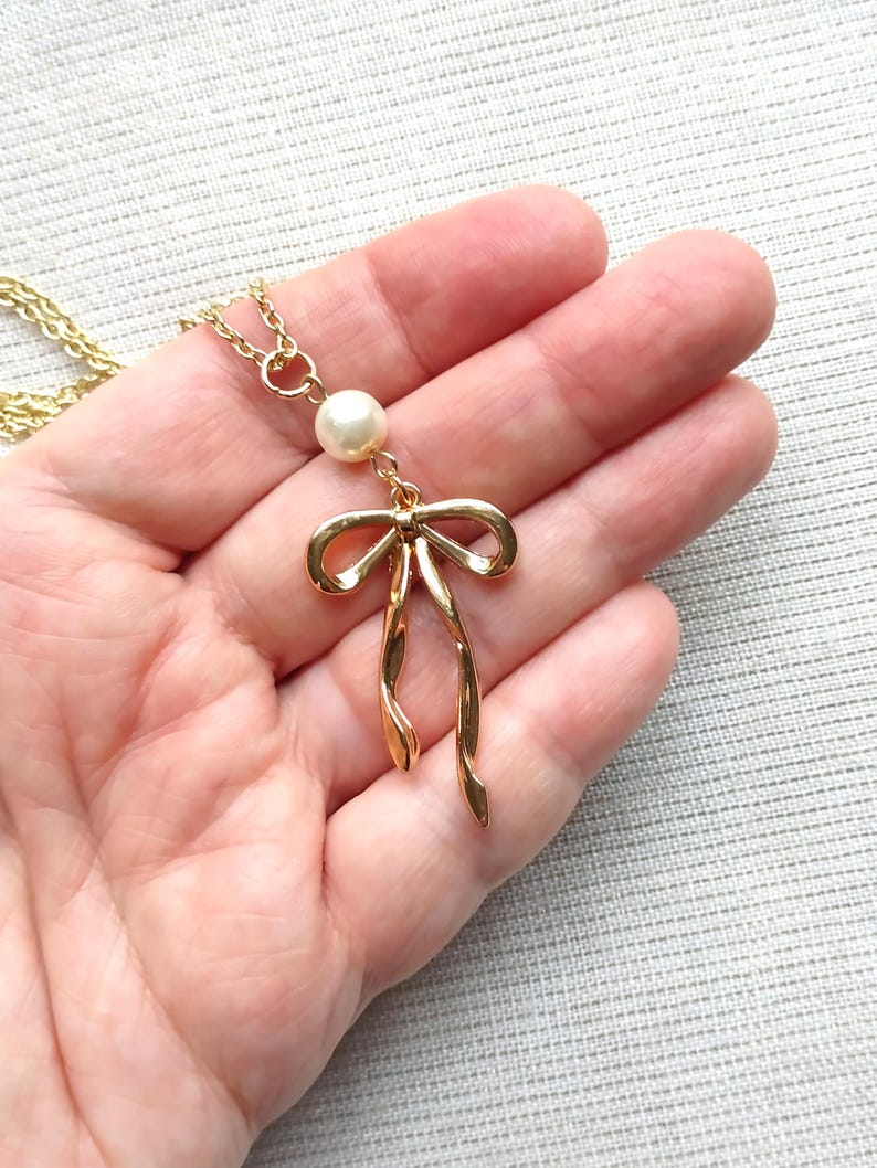 May include: A gold-colored necklace featuring a bow pendant with a dangling design. The necklace includes a small, round, white pearl above the bow. The chain is also gold-colored.