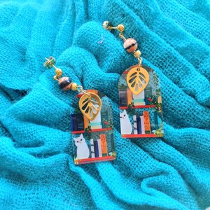 May include: A pair of dangle earrings featuring a whimsical design. Each earring has a gold-colored leaf charm, a rectangular pendant with a cat illustration, and decorative beads. The earrings are set against a turquoise fabric.