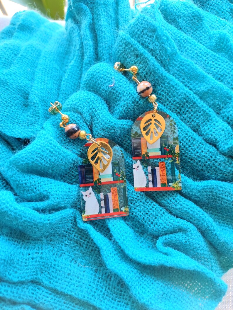 May include: A pair of dangle earrings featuring a whimsical design. Each earring has a gold-colored leaf charm, a rectangular pendant with a cat illustration, and decorative beads. The earrings are set against a turquoise fabric.