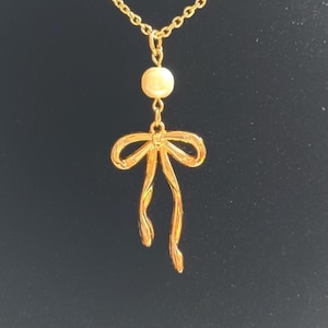 May include: A gold-colored necklace with a bow pendant and a small, round, off-white bead. The bow pendant has flowing ribbons. The necklace chain is delicate and gold-toned, set against a dark background.