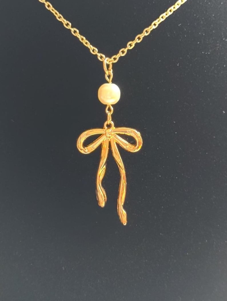 May include: A gold-colored necklace with a bow pendant and a small, round, off-white bead. The bow pendant has flowing ribbons. The necklace chain is delicate and gold-toned, set against a dark background.
