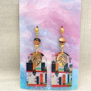 May include: A pair of dangle earrings featuring a gold-colored leaf charm, a marbled bead, and a gold-colored bead. The earrings have a clear, arched design with a cat, books, and greenery.