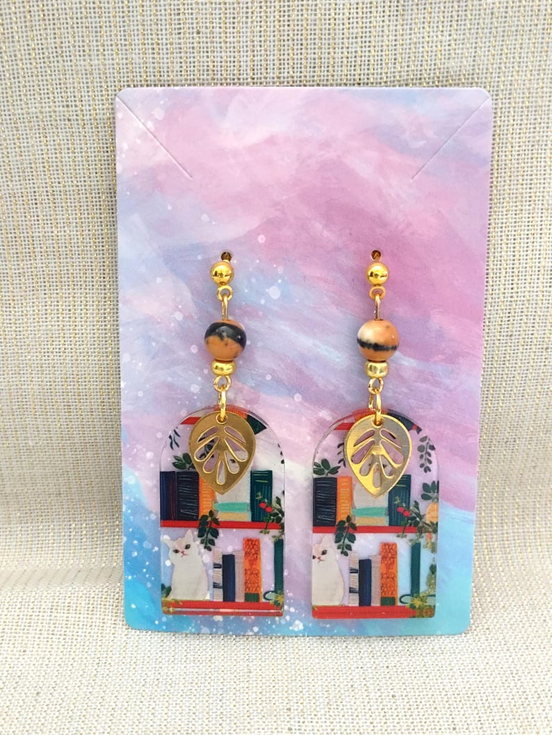 May include: A pair of dangle earrings featuring a gold-colored leaf charm, a marbled bead, and a gold-colored bead. The earrings have a clear, arched design with a cat, books, and greenery.