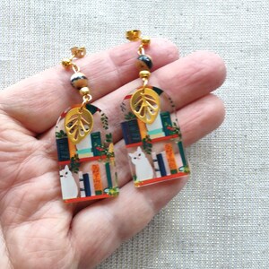 May include: A pair of dangle earrings featuring a whimsical design. Each earring showcases a miniature bookshelf scene with a white cat, books, and greenery. Gold-colored leaf charms and beads add detail. The earrings are held in a hand.