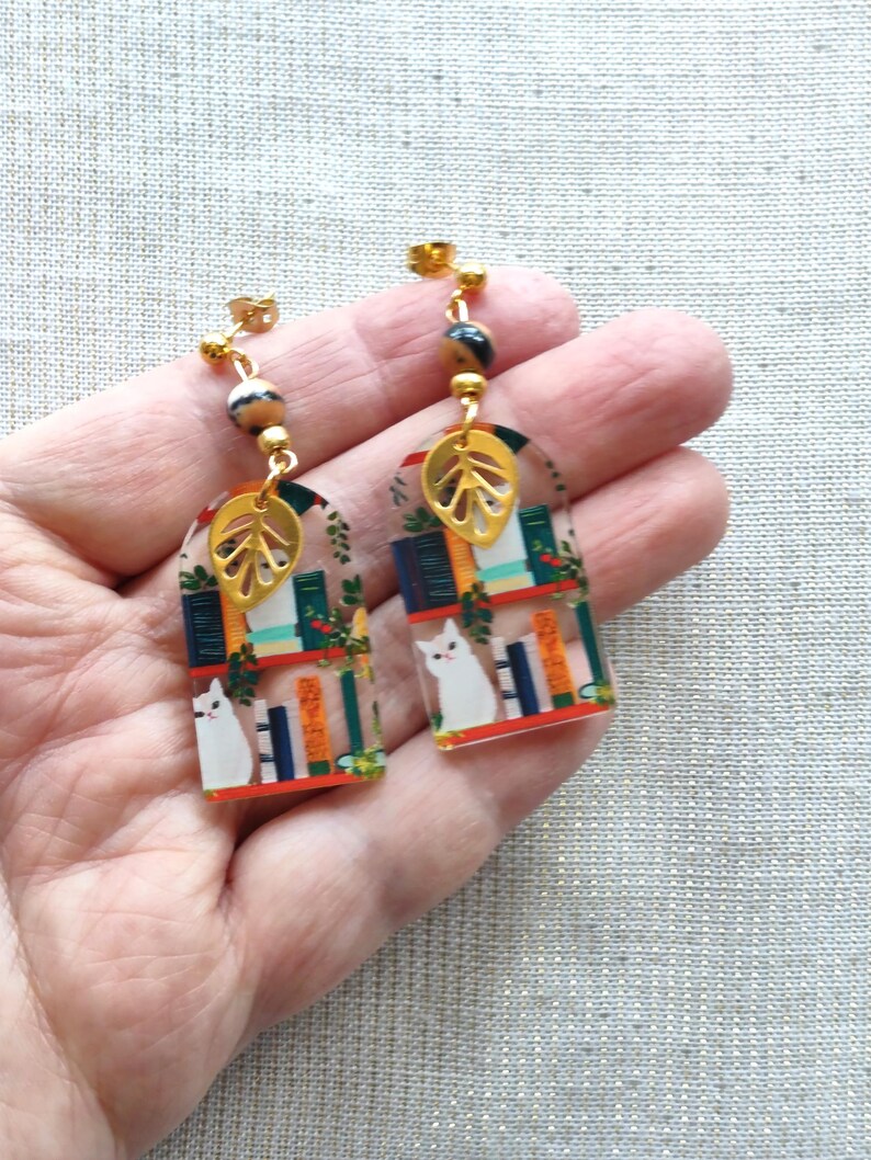 May include: A pair of dangle earrings featuring a whimsical design. Each earring showcases a miniature bookshelf scene with a white cat, books, and greenery. Gold-colored leaf charms and beads add detail. The earrings are held in a hand.