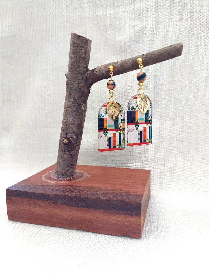 May include: A pair of colorful, patterned earrings with gold accents and leaf charms. The earrings hang from a wooden display stand. The earrings feature a mix of colors and geometric patterns, with a gold bead and leaf detail.
