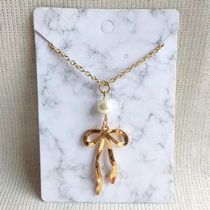 May include: Gold-toned necklace featuring a bow pendant with a dangling pearl. The necklace is displayed on a white marble-patterned card. The chain is delicate, and the bow has a polished finish.