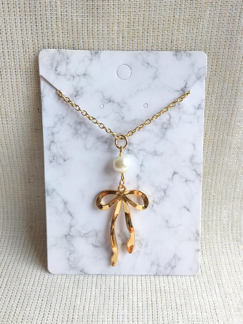 May include: Gold-toned necklace featuring a bow pendant with a dangling pearl. The necklace is displayed on a white marble-patterned card. The chain is delicate, and the bow has a polished finish.