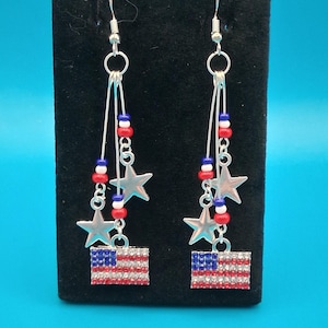 Pode incluir: Um par de brincos patrióticos com a bandeira americana. Cada brinco tem um gancho prateado, estrelas prateadas penduradas e um pequeno desenho da bandeira com contas vermelhas, brancas e azuis. O desenho da bandeira é feito de strass vermelhos e brancos.