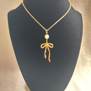 May include: A gold-colored necklace with a bow pendant and a small, round, off-white bead. The necklace is displayed on a black mannequin. The bow pendant has a delicate, open design.