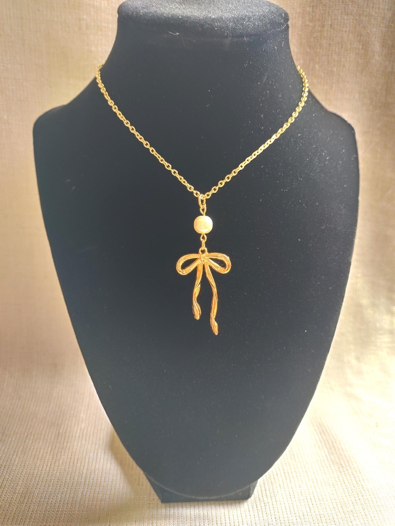 May include: A gold-colored necklace with a bow pendant and a small, round, off-white bead. The necklace is displayed on a black mannequin. The bow pendant has a delicate, open design.