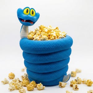 Pode incluir: Uma tigela de pipoca em forma de cobra azul, cheia de pipoca amarela. A cabeça da cobra, com olhos amarelos e boca vermelha, está no topo. Pedaços de pipoca estão espalhados ao redor da base da tigela.