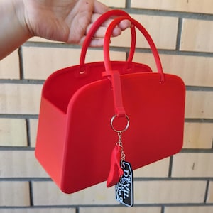 Puede incluir: Un bolso rojo brillante con dos asas redondeadas y forma rectangular. Una pequeña etiqueta con el texto "THE DEVIL WEARS ADA 2" cuelga de un anillo plateado sujeto al bolso. El bolso es sostenido por una mano.