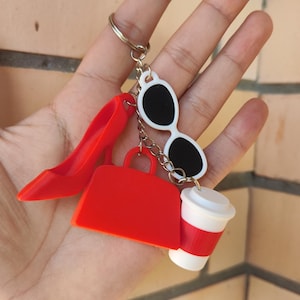 Könnte beinhalten: Ein Schlüsselanhänger mit Anhängern, darunter ein roter High-Heel-Schuh, eine rote Handtasche, eine weiße Sonnenbrille und eine Kaffeetasse. Die Anhänger sind an einem silbernen Schlüsselring befestigt. Der Hintergrund ist eine Backsteinmauer.