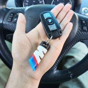 Op de afbeelding: Een zwarte BMW autosleutel met een zilverkleurig logo en een sleutelhanger in de vorm van de letter M, voorzien van een wit, blauw en rood gestreept ontwerp. De sleutel heeft drie knoppen en wordt vastgehouden voor een zwart lederen stuurwiel.