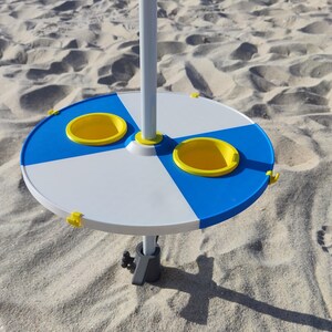 Pode incluir: Uma mesa de praia circular para fixar num poste, com um design segmentado em azul e branco, dois porta-copos amarelos e pequenos ganchos amarelos ao longo da borda externa, montada num poste central branco com uma base ajustável cinzento-escura.