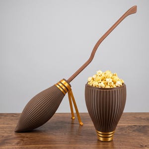 Può includere: Una scopa marrone con un lungo manico curvo e una base a coste a forma di goccia con bande dorate, accanto a una ciotola marrone abbinata, a coste e affusolata, riempita con una porzione di chicchi di popcorn gialli scoppiati.