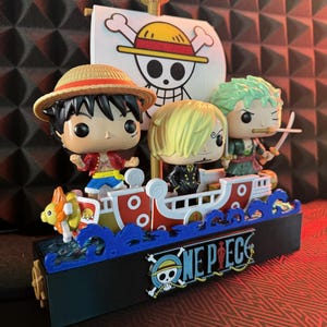 Può includere: Un oggetto da collezione di One Piece con tre personaggi su una nave. La nave è rossa e bianca con dettagli blu, e il logo di One Piece è mostrato sulla parte anteriore. Le figure includono Luffy, Zoro e Sanji.