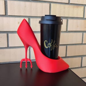 May include: A red high-heeled shoe-shaped coffee cup holder with a black travel mug. The travel mug has the word "Coffee" in gold script. The heel of the shoe has a trident design.