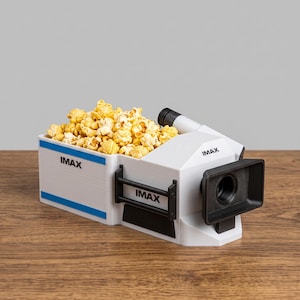 May include: A white plastic container shaped like a film projector filled with popcorn. The container features blue horizontal stripes and the black text IMAX printed on the side and front. It includes a black lens hood and a black railing detail.