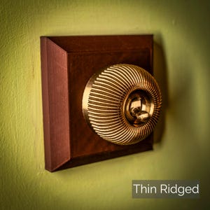 May include: A close-up of a gold-coloured light switch with a thin, ridged design. The switch is mounted in a dark brown rectangular plate. The text "Thin Ridged" is visible below the switch.