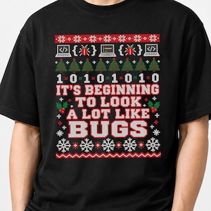 May include: A black short-sleeved t-shirt featuring a graphic print with a festive pattern of binary code, laptops, and Christmas trees. The text reads IT'S BEGINNING TO LOOK A LOT LIKE BUGS in white and red lettering, surrounded by snowflakes.