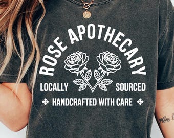 May include: Dark grey t-shirt with the text "ROSE APOTHECARY" in a curved arch, featuring two rose illustrations. The text "LOCALLY SOURCED HANDCRAFTED WITH CARE" is also printed on the shirt. The shirt is worn by a person.