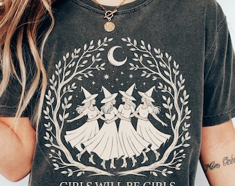 May include: Dark gray t-shirt featuring a cream-colored illustration of four witches dancing under a crescent moon and stars, framed by a leafy wreath. The text "GIRLS WILL BE GIRLS" is printed below the illustration. The shirt has a vintage, worn look.